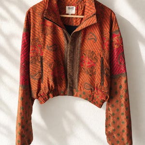 Op de afbeelding: Een kort jasje in oranje, bruine en rode tinten met een paisley- en bloemenpatroon. Het heeft een hoge kraag, een ritssluiting aan de voorzijde, lange mouwen met elastische boorden en een elastische zoom. Het binnenlabel toont de tekst "कला".