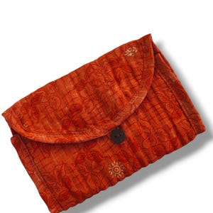Pode incluir: Uma bolsa retangular acolchoada de cor laranja com um padrão repetitivo de flores e grelha. A bolsa tem um fecho de aba curvo fixado por um único botão castanho escuro centrado na parte da frente do objeto.