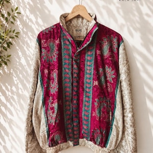 Puede incluir: Una chaqueta con cremallera que presenta un panel central magenta con patrones de cachemira en verde azulado y dorado, flanqueado por mangas y una espalda beige con un sutil estampado floral. La chaqueta tiene cuello alto, dobladillo elástico y etiqueta "कला KALA SARI".