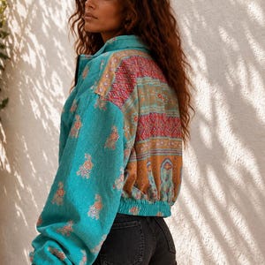 Puede incluir: Una chaqueta turquesa de manga larga con un panel trasero de patrones multicolores vibrantes en rojo, naranja y dorado con motivos geométricos y florales, combinada con pantalones cortos de mezclilla negros. La chaqueta tiene cuello alto y dobladillo elástico.