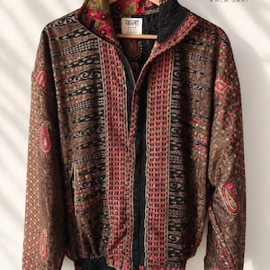 Op de afbeelding: Een bruin, zwart en roze jack met rits, een hoge kraag, lange mouwen en geribbelde boorden. De stof is voorzien van ingewikkelde paisley- en geometrische motieven. Een label in de nek bevat de tekst "कला KALA SARI".