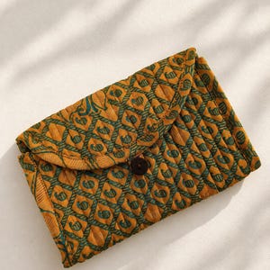 Op de afbeelding: Een gewatteerd rechthoekig tasje met een mosterdgele basis en een herhalend donkergroen paisley- en ruitpatroon. De envelopklep is aan de voorzijde bevestigd met een enkele donkerbruine, ronde knoop in het midden van de tas.