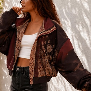 Puede incluir: Una chaqueta con cremallera que presenta una paleta de colores marrón oscuro y granate con intrincados patrones de cachemira en tonos dorados. La prenda tiene cuello alto, mangas largas con puños elásticos y un corte corto. El texto "कला KALA SARI" es visible.