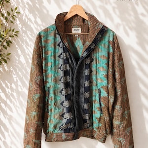 Puede incluir: Una chaqueta con cremallera que combina paneles de tela marrón y azul turquesa con patrones intrincados. La prenda incluye un cuello alto, mangas largas con puños elásticos y una banda vertical oscura a lo largo de la cremallera. Etiqueta KALA SARI.
