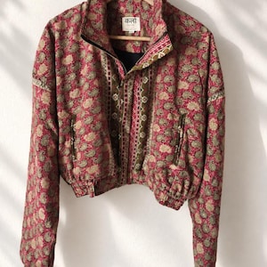 Può includere: Una giacca corta con motivo paisley rosso e verde, collo alto e chiusura frontale con zip. Il capo presenta pannelli decorativi verticali lungo la zip e polsini ed orlo elasticizzati. L'etichetta interna riporta la scritta "कला" e "KALA SARI".