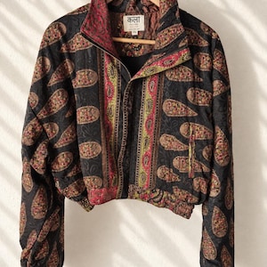 Op de afbeelding: Een kort jasje met een zwarte basis en een herhalend paisley-patroon in rood, groen en goud. Het jasje heeft een hoge kraag, een ritssluiting aan de voorzijde en elastische boorden en zoom. Het binnenlabel bevat de tekst "कला KALA SARI".