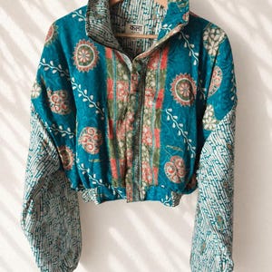 Op de afbeelding: Een kort jasje met een blauwgroene basis en een veelkleurig bloemen- en geometrisch patroon. Het kledingstuk heeft lange mouwen met een wit en blauwgroen baksteenachtig motief, een hoge kraag en een knoopsluiting aan de voorzijde. Het label toont "कला".