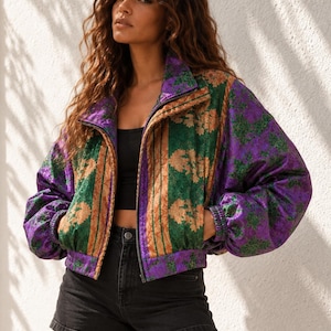 Könnte beinhalten: Eine Bomberjacke mit Reißverschluss und einem lebendigen Muster aus violetten und grünen Blumenmotiven mit vertikalen beigen Streifen. Die Jacke hat einen Stehkragen, lange Ärmel mit elastischen Bündchen und einen kurzen Saum, getragen über einem schwarzen Oberteil und Shorts.