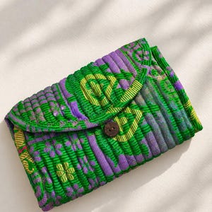 Könnte beinhalten: Eine gesteppte Stofftasche mit einem lebendigen grünen und violetten Blumenmuster und einer strukturierten, gerippten Oberfläche. Das rechteckige Accessoire verfügt über eine Umschlagklappe, die mit einem einzelnen, mittigen, dunkelbraunen runden Holzknopf verschlossen wird.