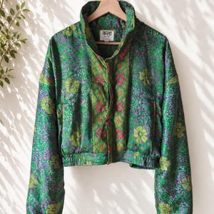 Pode incluir: Um casaco com estampa floral verde e roxa, gola alta, mangas compridas e bainha elástica. A frente apresenta um painel vertical central com um padrão geométrico em verde e rosa. Uma etiqueta interna exibe "कला" e "KALA SARI".