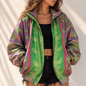 May include: A zip-up jacket featuring a vibrant patchwork design with green, purple, and pink hues. The garment displays a mix of paisley and leaf patterns, a high collar, long sleeves with elastic cuffs, and two side pockets, worn over a black top.