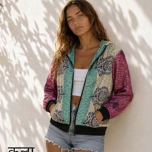 Puede incluir: Una chaqueta con capucha y cremallera que presenta un panel central de color crema con motivos de cachemira azul oscuro, flanqueado por bordes con patrón de chevrón turquesa y mangas magenta con un sutil estampado floral y puños negros.