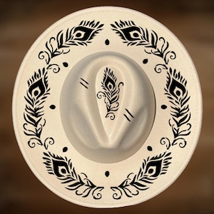 May include: Cream-colored fedora hat with a black peacock feather design around the brim and on the crown. The hat has a classic teardrop crown and a wide brim, perfect for sun protection. The hat is a stylish accessory.
