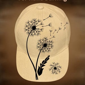 May include: A beige baseball cap with a black dandelion design. The cap features the text "For Hand Burning" and "CoolMoonDesigns". The dandelion design includes a large flower, seeds, and a leaf.