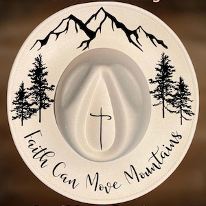 May include: A cream-colored fedora hat with a black mountain range and pine tree design. The hat features the text "Faith Can Move Mountains" and "For Hand Burning". The hat also has a cross design.