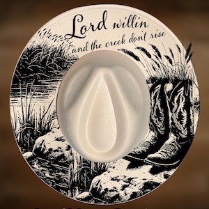 Puede incluir: Sombrero de vaquero color crema con una ilustración en blanco y negro de una escena de arroyo y botas de vaquero. El sombrero presenta el texto "Lord willin' and the creek don't rise" y el nombre del vendedor, CoolMoonDesigns.