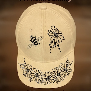 May include: A beige baseball cap featuring black line art of a bee, a single flower, and a floral garland on the brim. The text "For Hand Burning" appears at the top, and "CoolMoonDesigns" is printed at the bottom of the cap design.