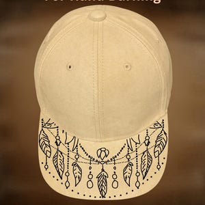 Könnte beinhalten: Eine beige Baseballkappe mit einem schwarzen Linienmuster auf dem Schirm, das Federn und Perlenschnüre darstellt. Der Text "For Hand Burning" steht über der Kappe und "CoolMoonDesigns" ist in weißer Schrift unter dem Schirm zu sehen.