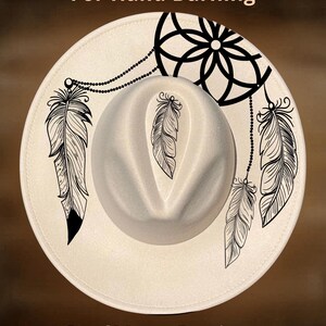 Puede incluir: Sombrero fedora color crema con diseños de plumas y atrapasueños negros. El sombrero presenta el texto "For Hand Burning" y "CoolMoonDesigns". Un accesorio elegante.