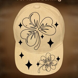 May include: A beige baseball cap featuring black line art of two shamrocks and several four-pointed stars. The text "For Hand Burning" is printed above the design, and "CoolMoonDesigns" is printed below the design in a simple font.