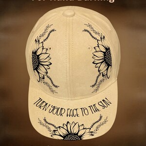 May include: Cream-colored baseball cap with black sunflower and leaf designs. The text "TURN YOUR FACE TO THE SUN" is on the brim. The words "For Hand Burning" and "CoolMoonDesigns" are also visible.