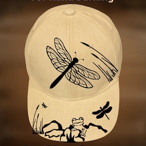 May include: A beige baseball cap with a black dragonfly design and the text "For Hand Burning" and "CoolMoonDesigns". The cap features a dragonfly, frog, and cattails design.