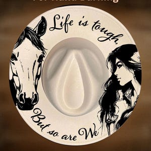 May include: A cream-colored fedora hat featuring a black graphic illustration of a horse on the left and a woman in profile on the right. The text "Life is tough, But so are We" is printed in black script. The hat is labeled For Hand Burning.