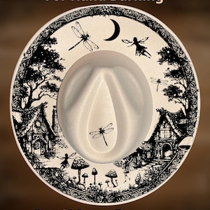 Puede incluir: Un sombrero fedora color crema con una detallada ilustración negra alrededor del borde. El diseño incluye libélulas, hadas, una luna creciente y casas caprichosas. También se ve el texto "For Hand Burning" y "CoolMoonDesigns".