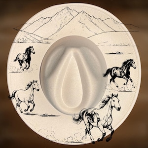 Puede incluir: Sombrero de vaquero color crema con ilustraciones de montañas y caballos corriendo. El sombrero presenta el texto "For Hand Burning" y "CoolMoonDesigns". El ala está decorada con cuatro caballos corriendo.
