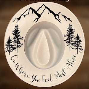 Może przedstawiać: Kremowy kapelusz fedora z motywem gór i drzew. Kapelusz ma napis "For Hand Burning" na górze i "Go Where You Feel Most Alive" na rondzie. Nazwa marki "CoolMoonDesigns" znajduje się na dole.