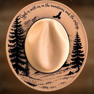 May include: Tan fedora hat with a cream-colored crown. The brim features a mountain and tree design with the text "God is with us in the mountains and the valleys." The text "For Hand Burning" and "CoolMoonDesigns" are also visible.