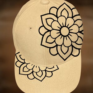 May include: A beige baseball cap featuring a black floral design on the front and bill. The cap has a curved bill and a button on top. The floral design is a stylized mandala.