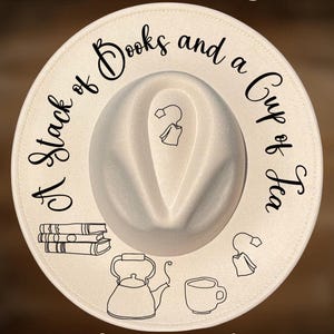 Könnte beinhalten: Cremefarbener Fedora-Hut mit dem Text "A Stack of Books and a Cup of Tea" in Schreibschrift. Der Hut zeigt Illustrationen von Büchern, einer Teekanne, einer Teetasse und Teebeuteln. Der Text "For Hand Burning" und "CoolMoonDesigns" sind ebenfalls vorhanden.