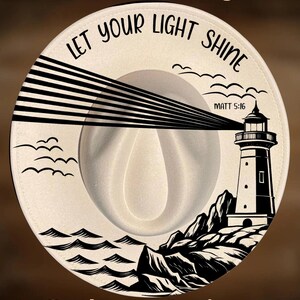 May include: A cream-colored fedora hat featuring a black graphic of a lighthouse on a rocky shore with waves and birds. The text reads "For Hand Burning", "LET YOUR LIGHT SHINE", "MATT 5:16", and "CoolMoonDesigns" in black lettering.