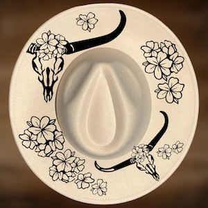 May include: Cream-colored fedora hat with a wide brim. The hat features black floral and bull skull designs around the crown. The hat is a stylish accessory for various occasions.