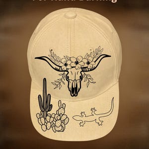 Può includere: Cappellino da baseball beige con un design a teschio di toro nero, accenti floreali e illustrazioni a tema desertico. È visibile anche il testo "For Hand Burning" e "CoolMoonDesigns".