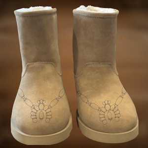May include: A pair of beige suede boots with a decorative black design on the front. The boots have a round toe, a short shaft, and a faux fur lining. The soles are a lighter beige color.