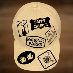 May include: A cream-coloured baseball cap featuring several black graphic patches. Patches include the text "Happy Camper" and "National Parks", a compass, bear paw prints, a cactus, and a mountain scene. The text "For Hand Burning" and "CoolMoonDesigns" are also visible.