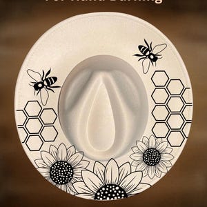 May include: A cream-coloured fedora hat featuring black line art illustrations of two bees, two honeycomb patterns, and three sunflowers. The text "For Hand Burning" appears at the top and "CoolMoonDesigns" is printed at the bottom.