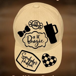 May include: A beige baseball cap featuring black graphic designs including a bow, a tumbler, a checkered heart, and text that reads Eye Candy, A lil' Bougie, and Somebody's Problem. The text For Hand Burning and CoolMoonDesigns is visible.