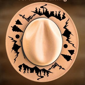 Puede incluir: Sombrero estilo fedora color beige con ala negra y corona crema. El sombrero presenta un diseño decorativo negro de montañas y árboles alrededor del ala. También se ve el texto "For Hand Burning" y "CoolMoonDesigns".