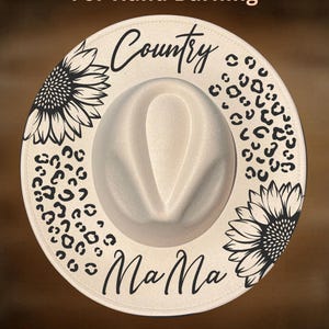 Könnte beinhalten: Cremefarbener Fedora-Hut mit breiter Krempe. Der Rand hat schwarze Sonnenblumen- und Leopardenmuster mit den Worten "Country Mama" in Schrift. Der Text "For Hand Burning" und "CoolMoonDesigns" sind ebenfalls sichtbar.