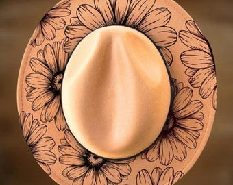Puede incluir: Un sombrero fedora beige con una corona marrón claro y un ala ancha. El ala está decorada con un patrón repetido de grandes flores negras estilizadas. El texto "For Hand Burning" y "CoolMoonDesigns" está impreso en el sombrero.