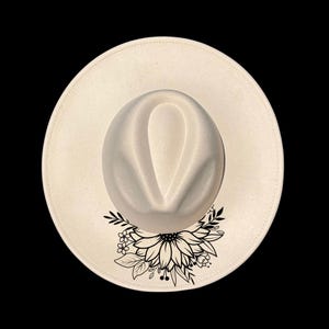 May include: Cream-colored fedora hat with a black floral design on the brim. The hat features a structured crown and a wide brim. The floral design includes a sunflower and leaves.