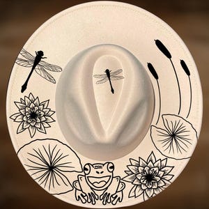 May include: Cream-colored fedora hat with a black dragonfly, frog, lily pad, and cattail design. The hat features a wide brim and a structured crown. The illustrations are detailed and create a nature-inspired theme.