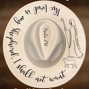 May include: Cream-colored fedora hat with a white brim. The hat features the text "The Lord is my shepherd. I shall not want" and "Psalm 23:1". A shepherd and sheep are illustrated on the brim. The text "For Hand Burning" and "CoolMoonDesigns" are also present.