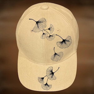 May include: A beige baseball cap with a curved brim. The cap features a black ginkgo leaf design and the text "For Hand Burning" and "CoolMoonDesigns".