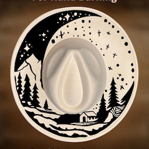 Può includere: Un cappello fedora color crema con un disegno nero bruciato a mano sul bordo. Il disegno presenta una catena montuosa, alberi, una casa e una luna crescente con stelle. È visibile anche il testo "For Hand Burning" e "CoolMoonDesigns".