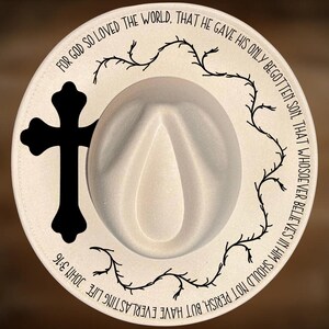 May include: A cream-colored cowboy hat with a black cross and a decorative vine design. The hat features the text "For God so loved the world, that he gave his only begotten son, that whosoever believes in him should not perish, but have everlasting life John 3:16." The text "For Hand Burning" and "CoolMoonDesigns" are also visible.