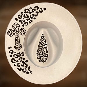 May include: Cream-colored fedora hat with a wide brim. The hat features a black cross design and leopard print accents on the brim and crown. The hat is a stylish accessory.